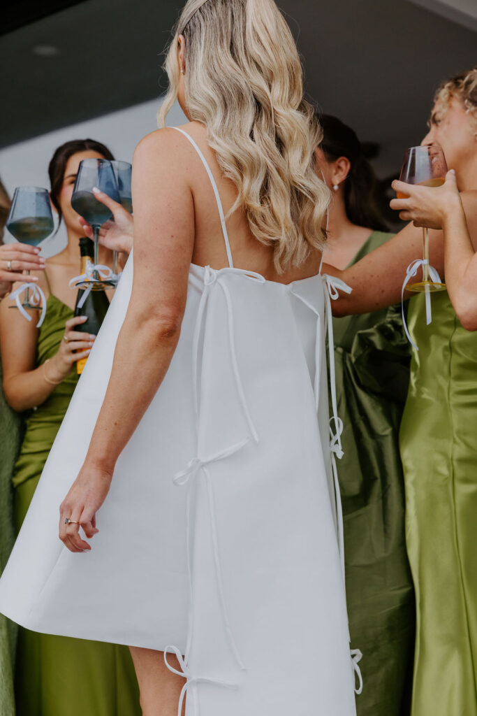 Back view of a bride in a white dress with side ties, surrounded by friends in olive dresses, toasting with wine glasses in celebration