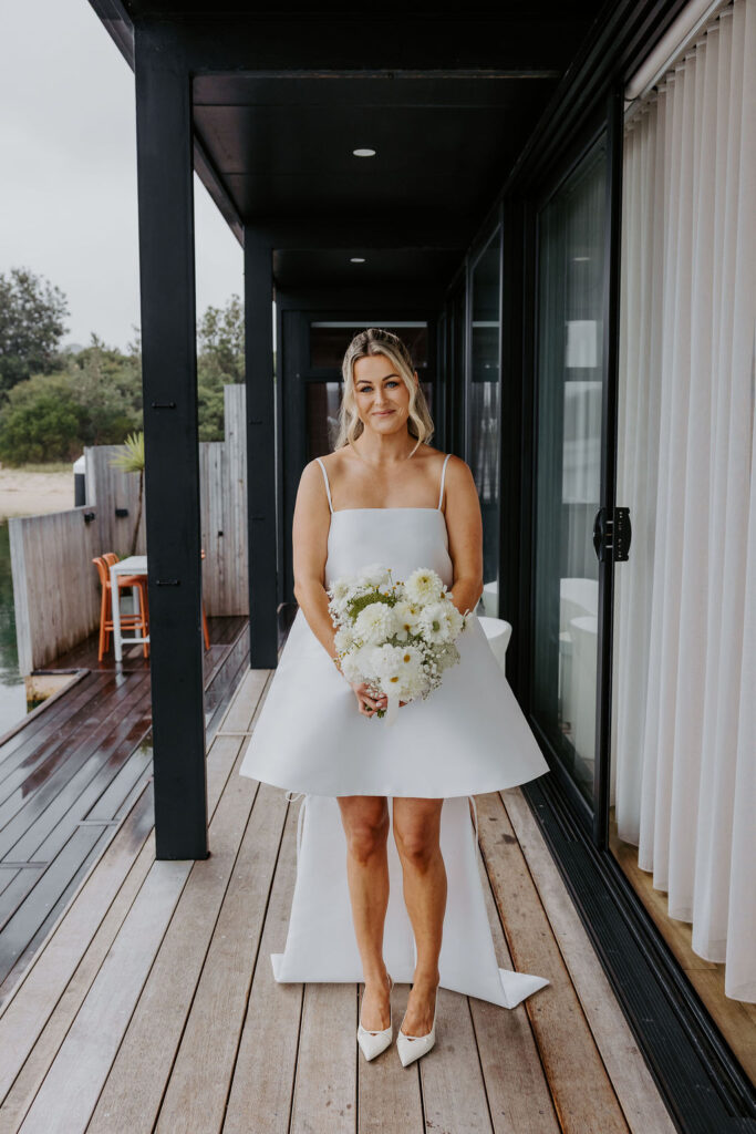 Bride in a short white strap-shoulder dress holding a white flower bouquet, standing on a wooden deck outside a modern building.
