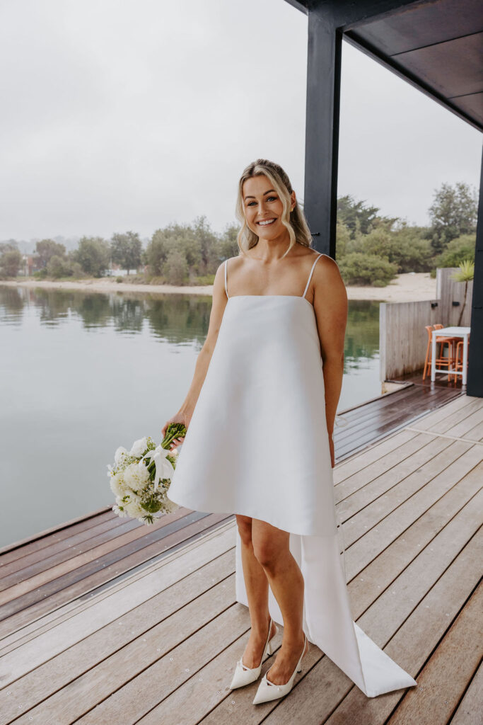 Bride standing on a wooden dock by a calm lake, holding a white bouquet and wearing a simple white wedding dress with thin straps.