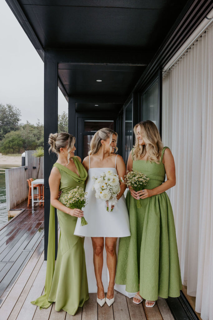 Bride in a white dress joined by two bridesmaids in green gowns, all holding bouquets on a wooden deck beside a glass-walled building.