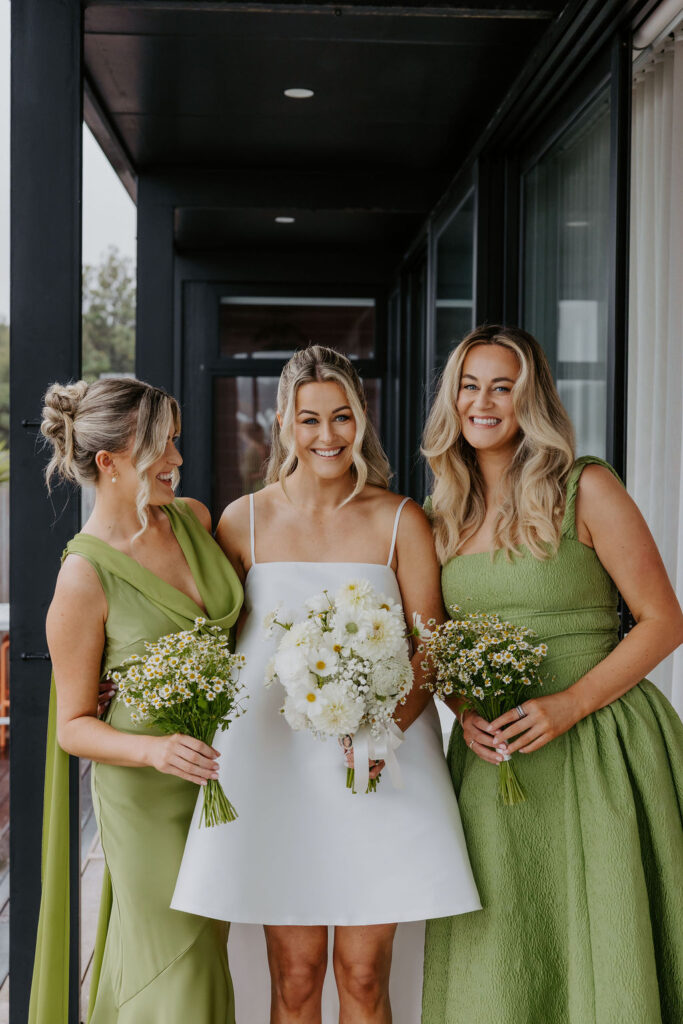 Three women in dresses smile for a wedding photo; the bride in white stands between two bridesmaids in light green, each holding bouquets of white flowers.