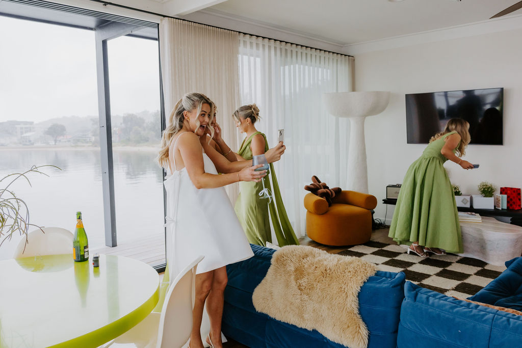 Bride and bridesmaids in a bright lakeside living room, taking a selfie; champagne on a lime table nearby.
