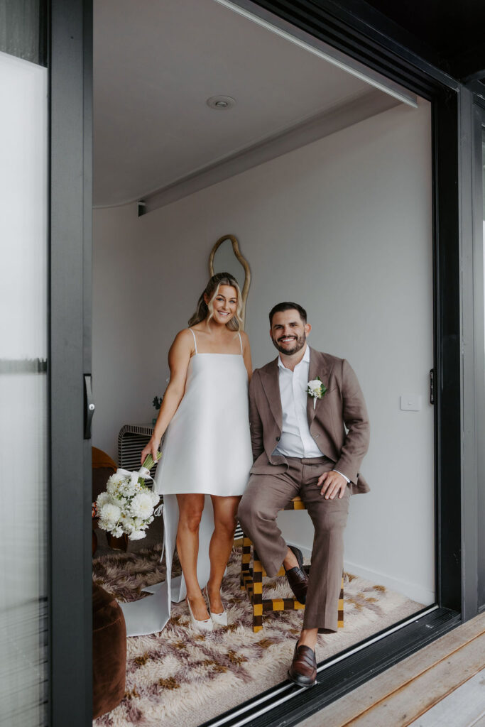 Bride in a white dress stands beside a groom in a brown suit inside a modern room, holding a white bouquet.
