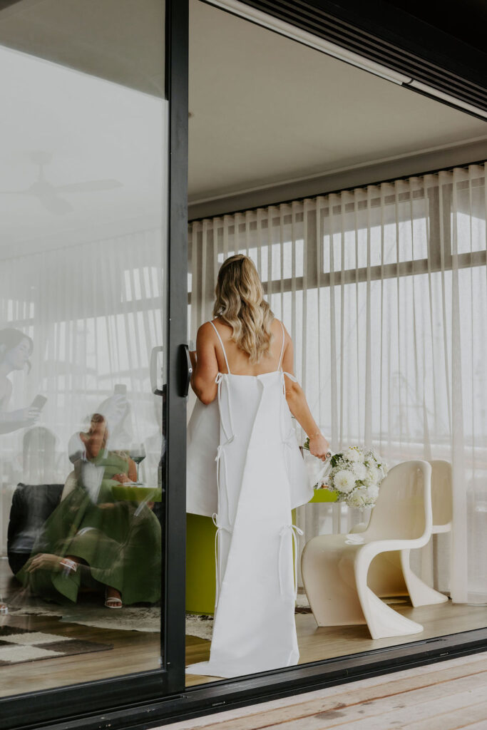 Bride in a white wedding gown seen from behind, holding a bouquet, standing inside a glass-walled room with sheer curtains.