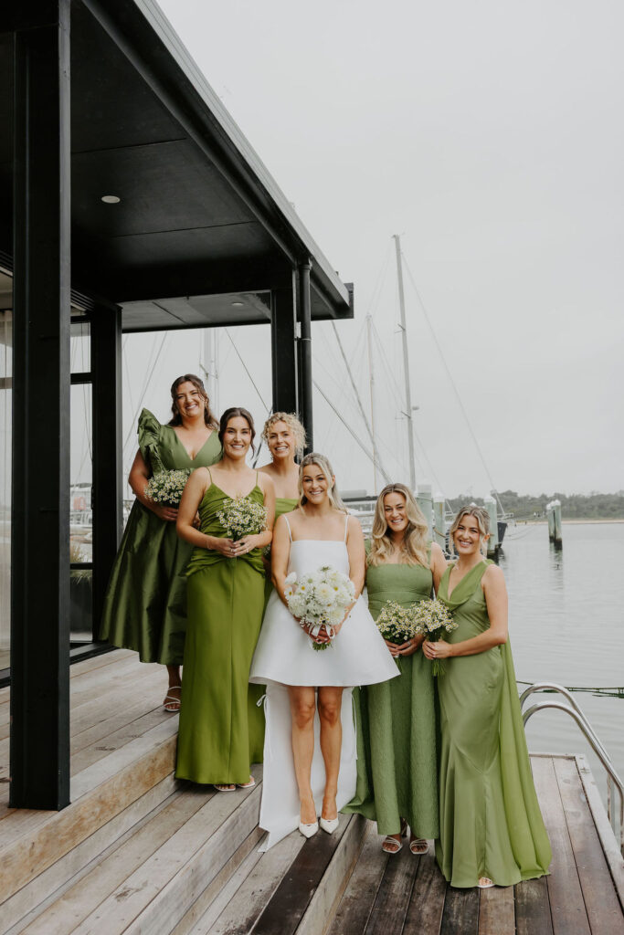 Bride in white dress holding bouquet with her bridesmaids in green gowns posing on a dock by a harbor. Notable: calm water and sailboats in background.