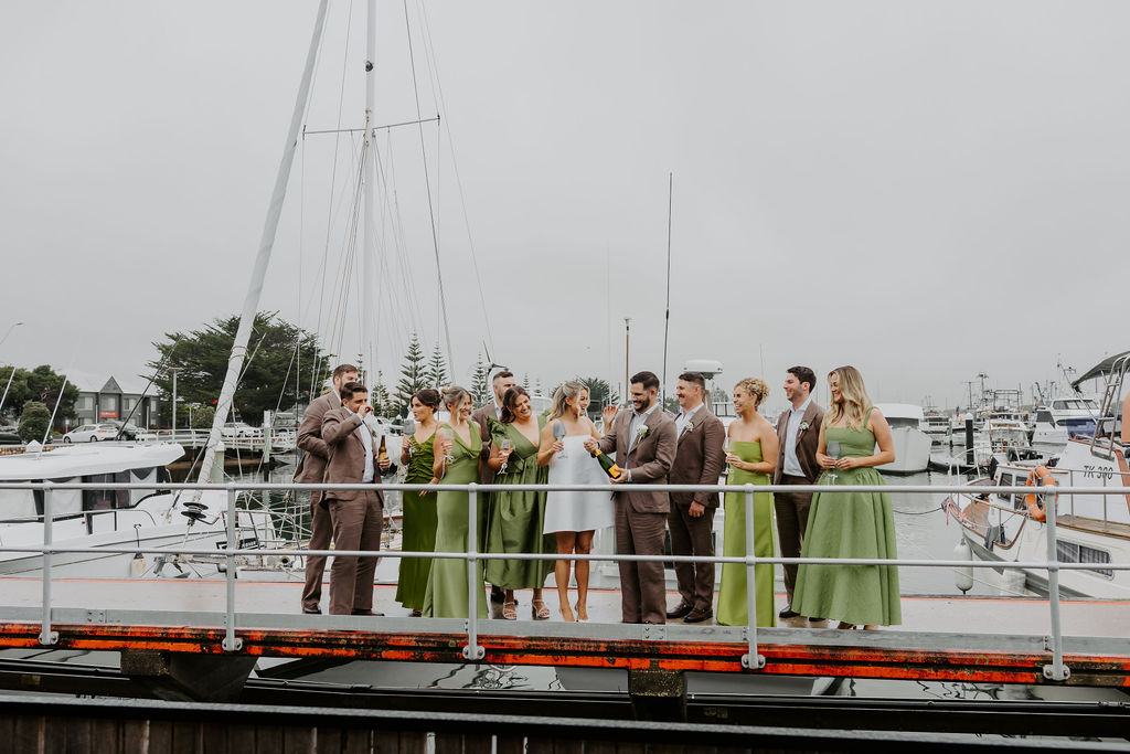 Wedding party on a marina pier, toasting with champagne as bridesmaids in green dresses and groomsmen in brown suits stand with the couple, sailboats in the background.
