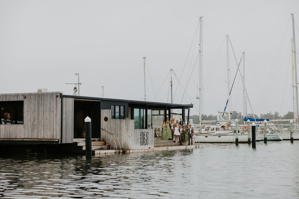 Group of people gathered on a wooden dock beside a modern marina building, with sailboats moored in the background.