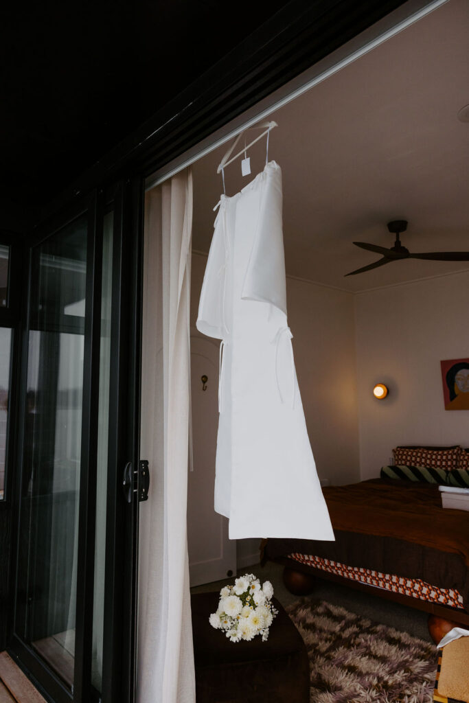 White wedding dress hanging on a hanger inside a bedroom near a glass door, with a bouquet on a dark ottoman nearby.