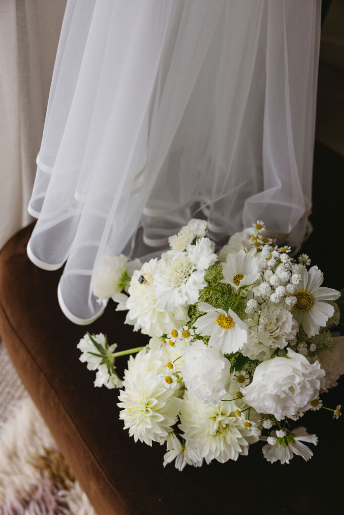 White wedding bouquet with daisies and hydrangea-like blooms resting on a dark chair, framed by a sheer veil.