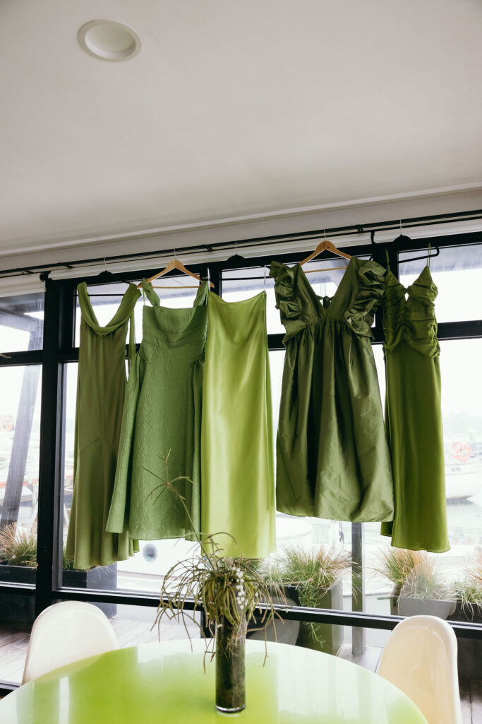Green dresses on hangers hanging from a rack along large windows, with a lime round table and white chairs in the foreground.
