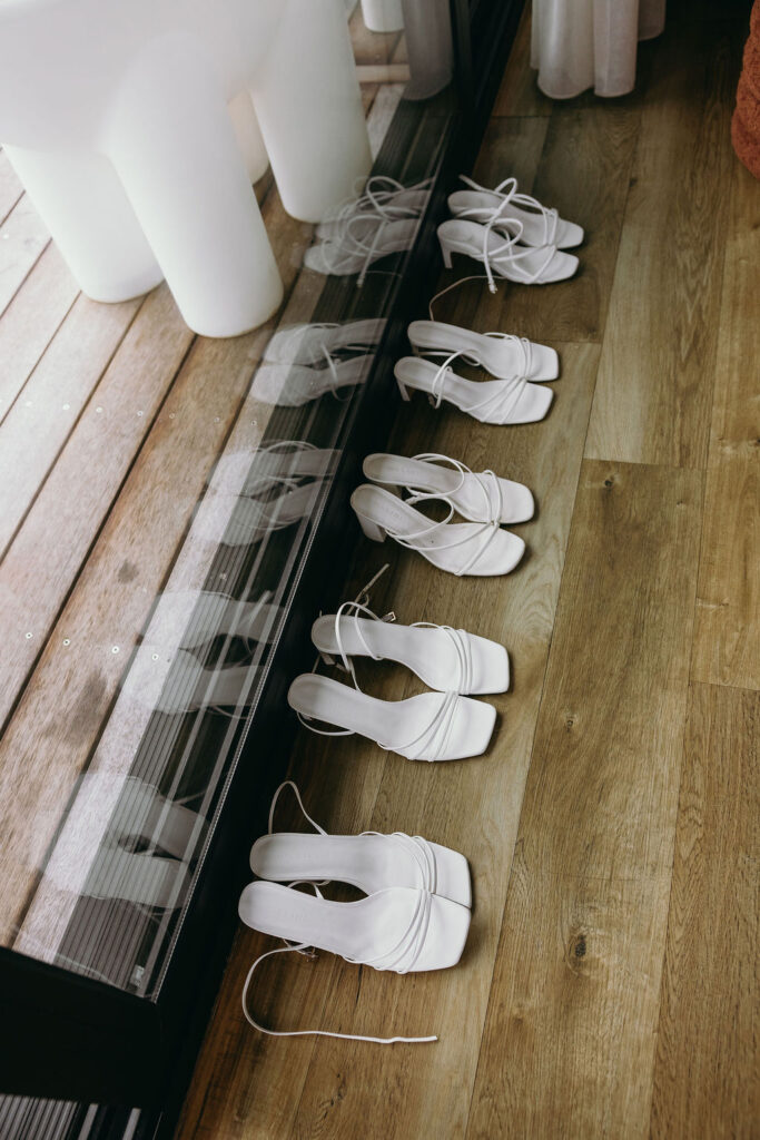 Row of white heeled sandals with thin straps lined up along a glass storefront panel on a wooden floor, showroom display.