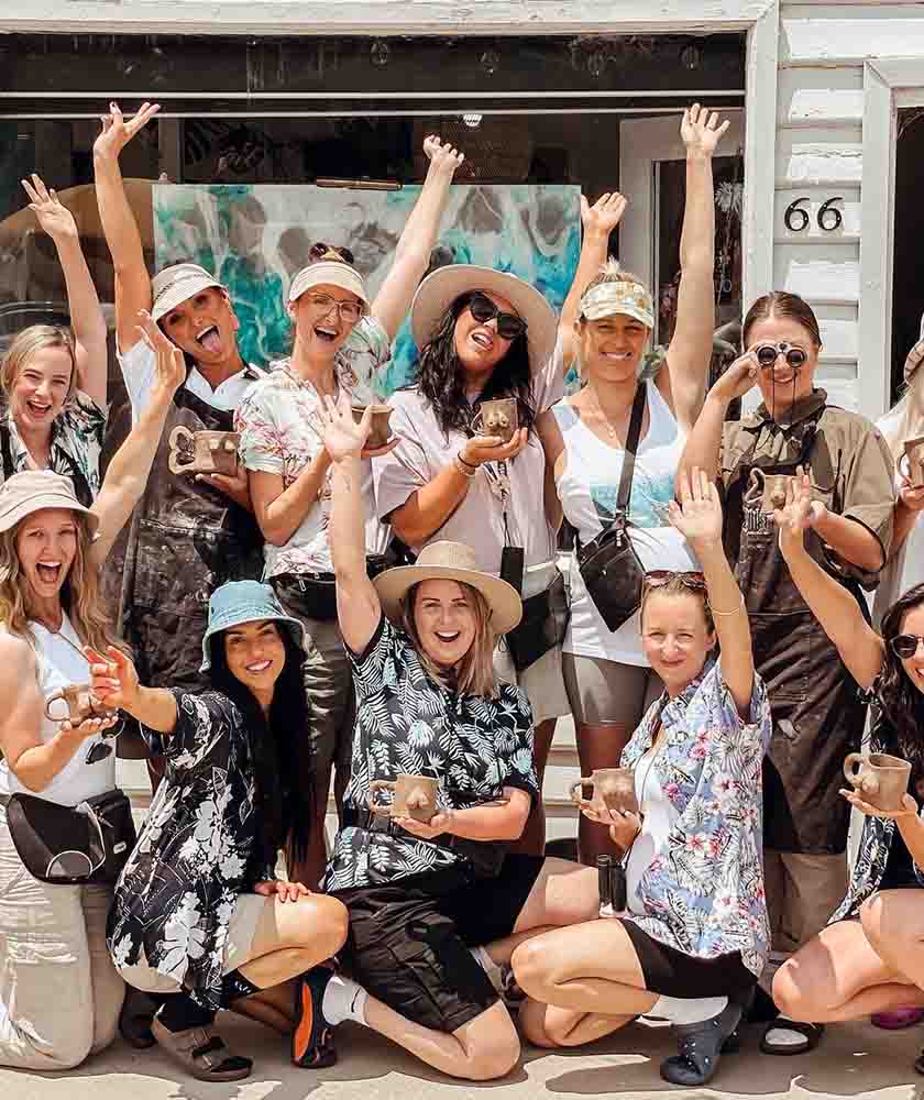 A cheerful group of people posing outdoors, holding mugs and raising arms in celebration in front of a storefront.