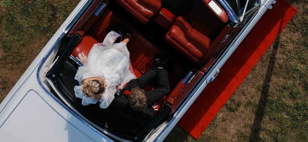 Top-down view of a bride in a white gown and a groom in a dark suit sitting in the back of a vintage car with red leather seats, on a red carpet outside.