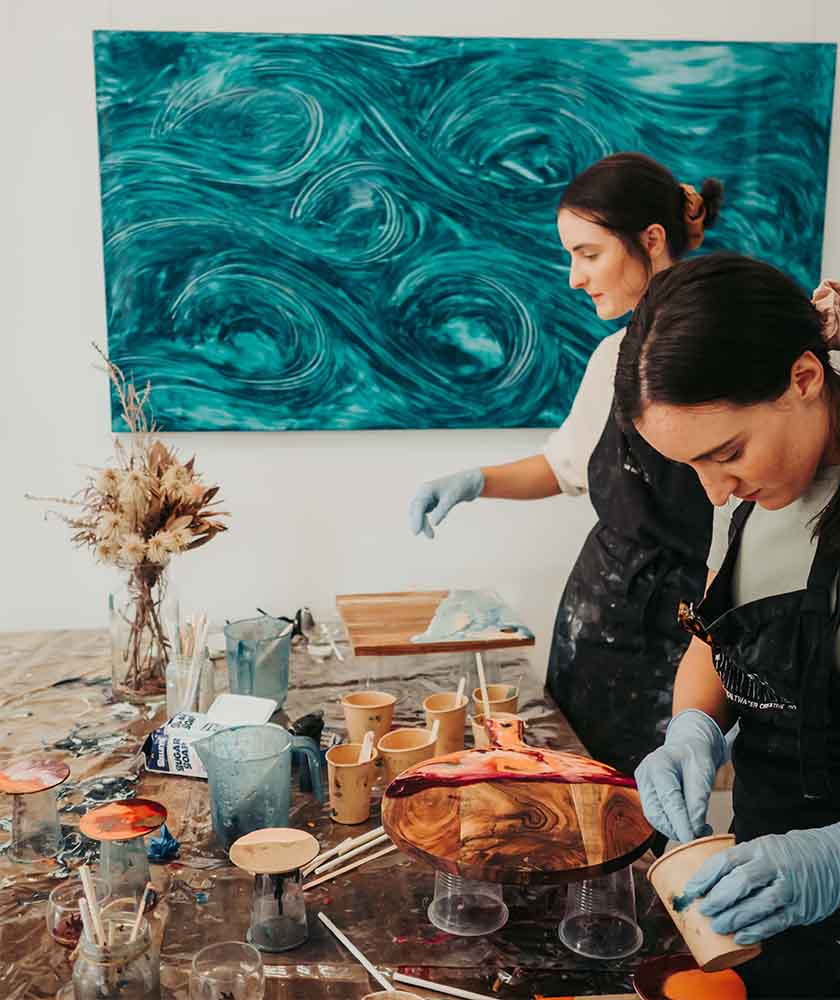 Two artists in aprons painting and sanding wooden pieces at a cluttered studio table with a teal swirled painting on the wall behind them