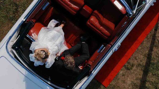 Top-down view of a bride in a white gown and a groom in a dark suit sitting in the back of a vintage car with red leather seats, on a red carpet outside.