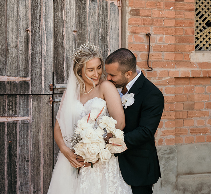 Bride in a lace wedding dress holds a white bouquet while a groom in a black suit affectionately embraces her outside a brick building with a wooden door, wedding day moment.
