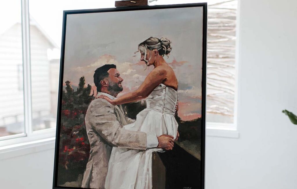 1 | I do I do Gippsland Framed painting showing a bearded man in a light suit embracing a woman in a white dress outdoors at sunset, on an easel inside a bright room.