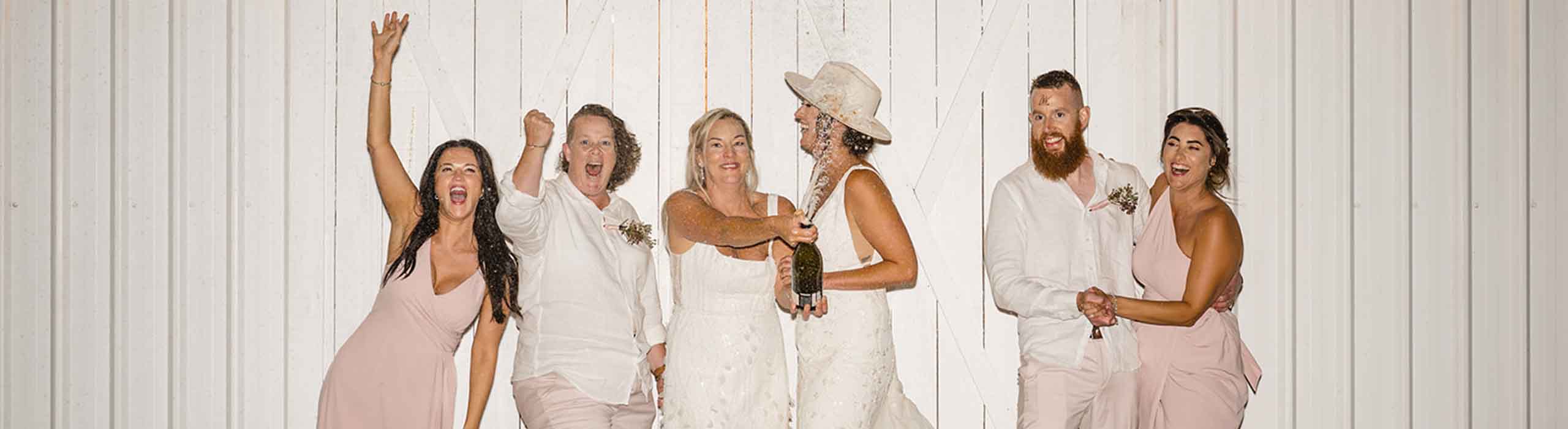 Wedding party celebrates: a bride in a white dress raises champagne with friends in light pink outfits against a white wooden wall.