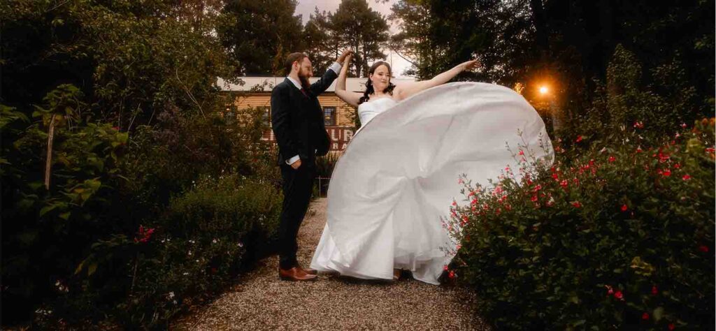 Bride in a white wedding gown twirls on a gravel path as her groom in a suit holds her hand in a garden setting at dusk.