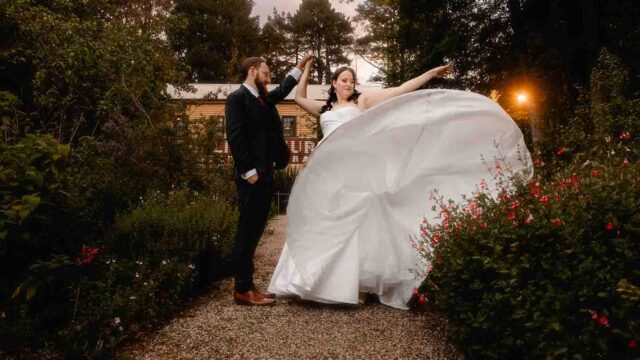 Bride in a white wedding gown twirls on a gravel path as her groom in a suit holds her hand in a garden setting at dusk.