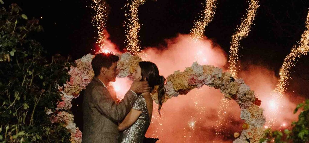 A couple kissing under a flower arch as bright fireworks explode behind them at night in a festive outdoor setting.