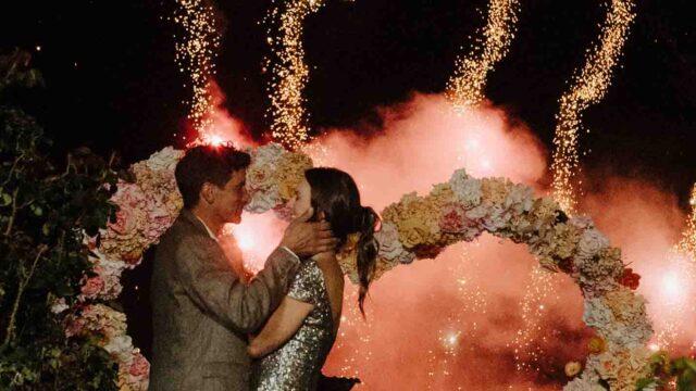 A couple kissing under a flower arch as bright fireworks explode behind them at night in a festive outdoor setting.