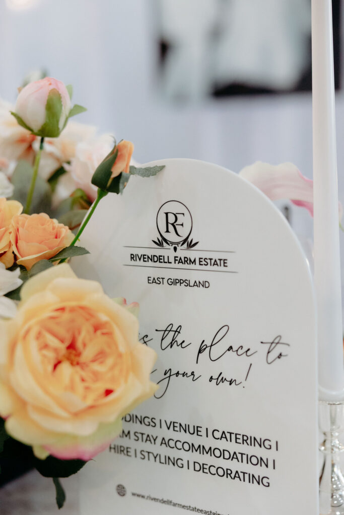 Peach and pink roses beside a white wedding invitation card with Rivendell Farm Estate logo.