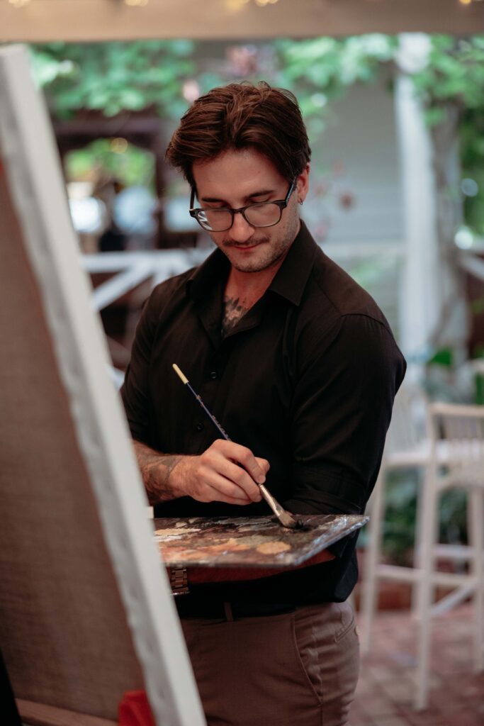 219A8675 3 | I do I do Gippsland Man wearing glasses and a black shirt paints on a wooden palette outdoors, focused on his work.