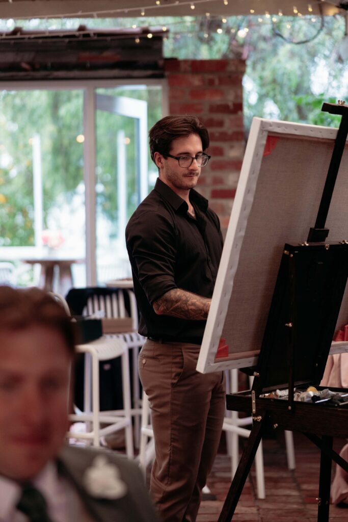 219A8697 1 | I do I do Gippsland Man with dark hair and glasses stands at an easel, painting on a canvas in a brick-walled room with string lights overhead.