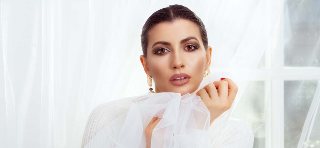Portrait of a woman with bold makeup holding sheer white fabric near her face in a bright, airy setting.