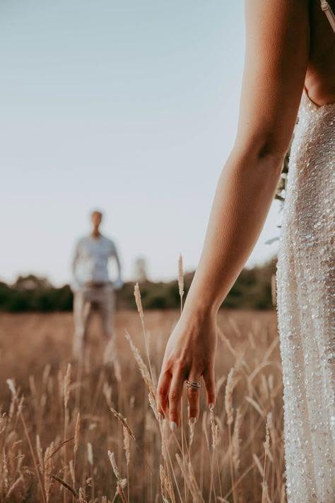 486067484_1068692971946587_1558429521068126386_n - I do I do Gippsland Bride’s beaded dress and hand with ring reaching toward tall grass, groom standing in the distance on a field.