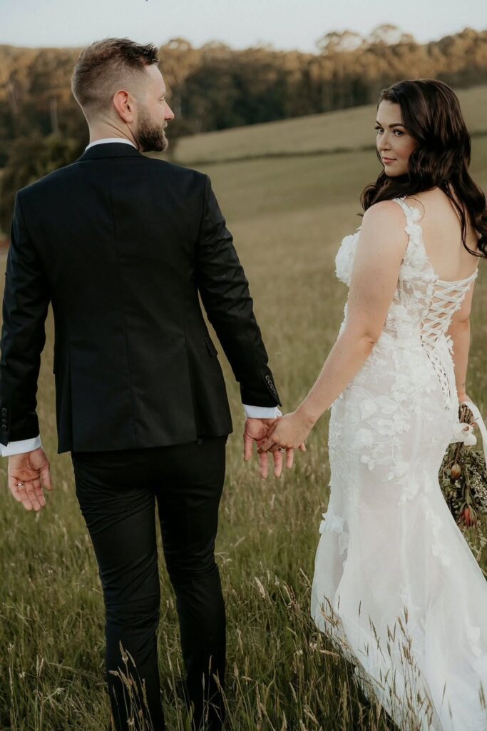 499824254_1119044726911411_1960323461094600445_n - I do I do Gippsland Bride and groom walk hand in hand through a grassy field, the bride in a lace wedding dress and the groom in a dark suit, seen from behind.