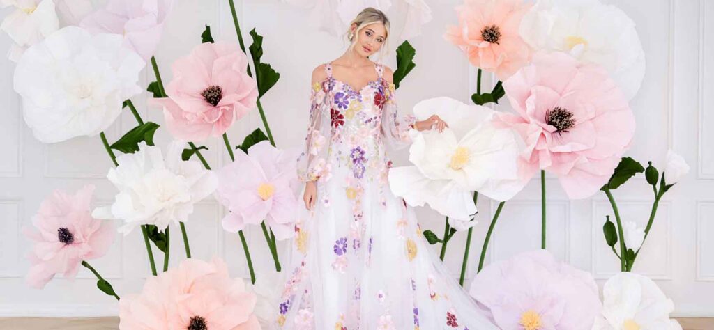 Woman in a long pastel floral gown posing among oversized pink and white paper flowers in a bright studio setting.