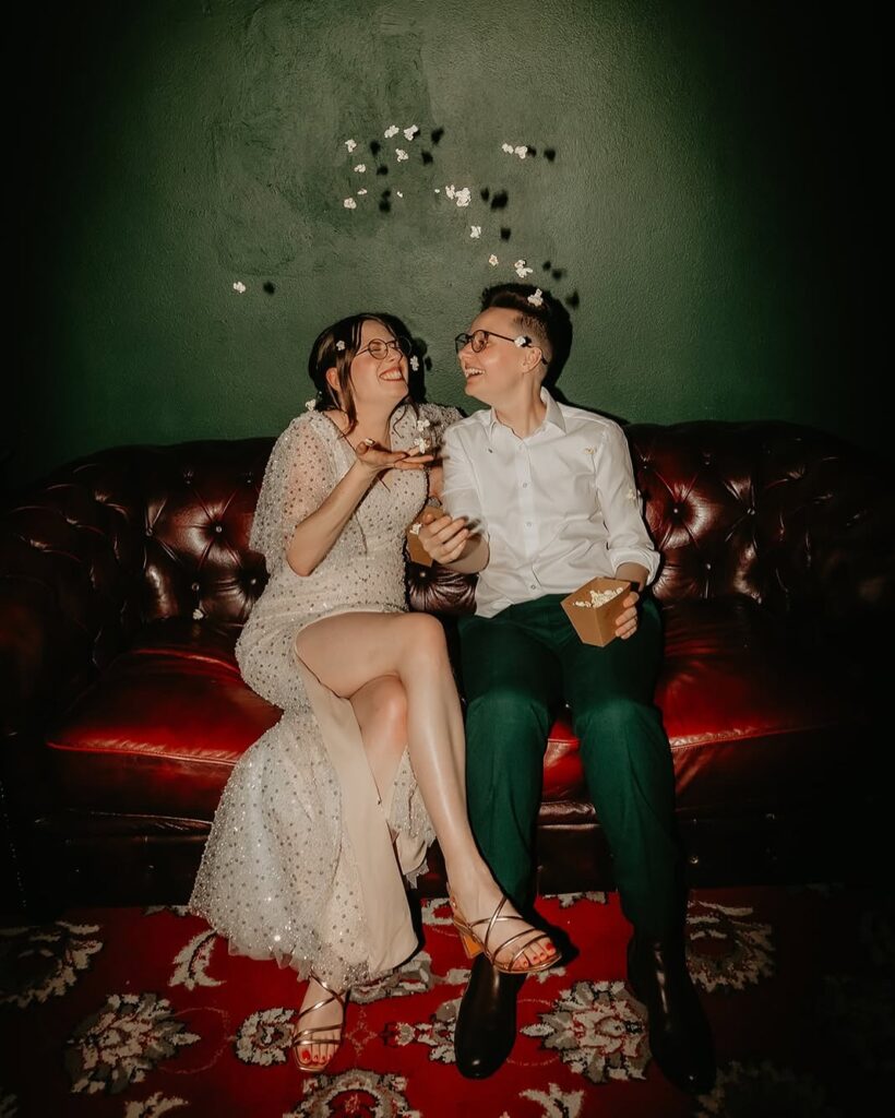 Had to be Weddings1 - I do I do Gippsland Two people sit close on a red leather couch, laughing as white confetti falls around them during a celebration.