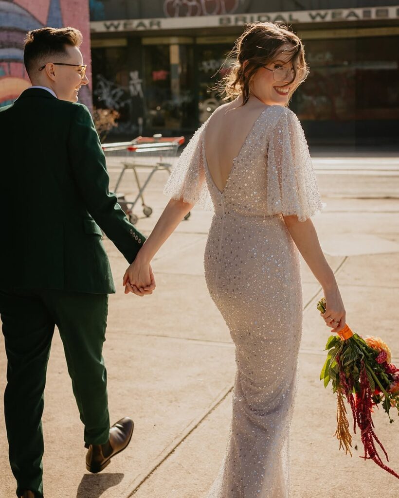 Had to be You Weddings2 - I do I do Gippsland Couple walking hand in hand on a city sidewalk; woman in a sparkly backless gown carrying a bouquet, man in a green suit beside her.