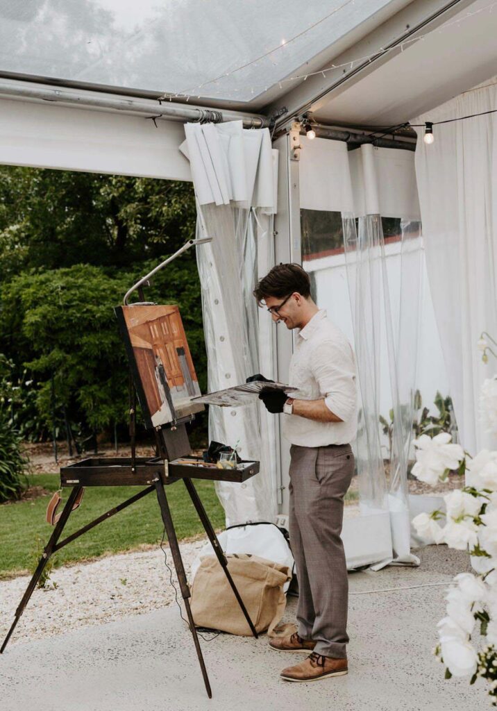 IMG_20250215_072319_862 | I do I do Gippsland Man in a light shirt and glasses paints at an outdoor easel inside a white-tented setup, holding a palette and brush.
