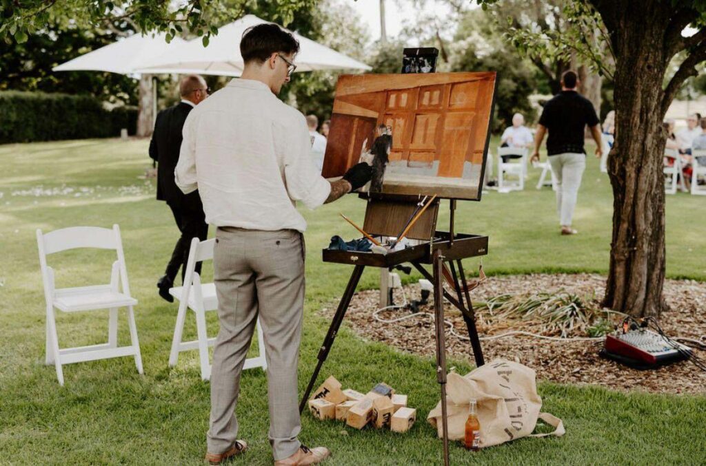 IMG_20250215_072326_046 | I do I do Gippsland Man in a white shirt and beige pants paints on an easel at an outdoor garden event, with white chairs and guests in the background.
