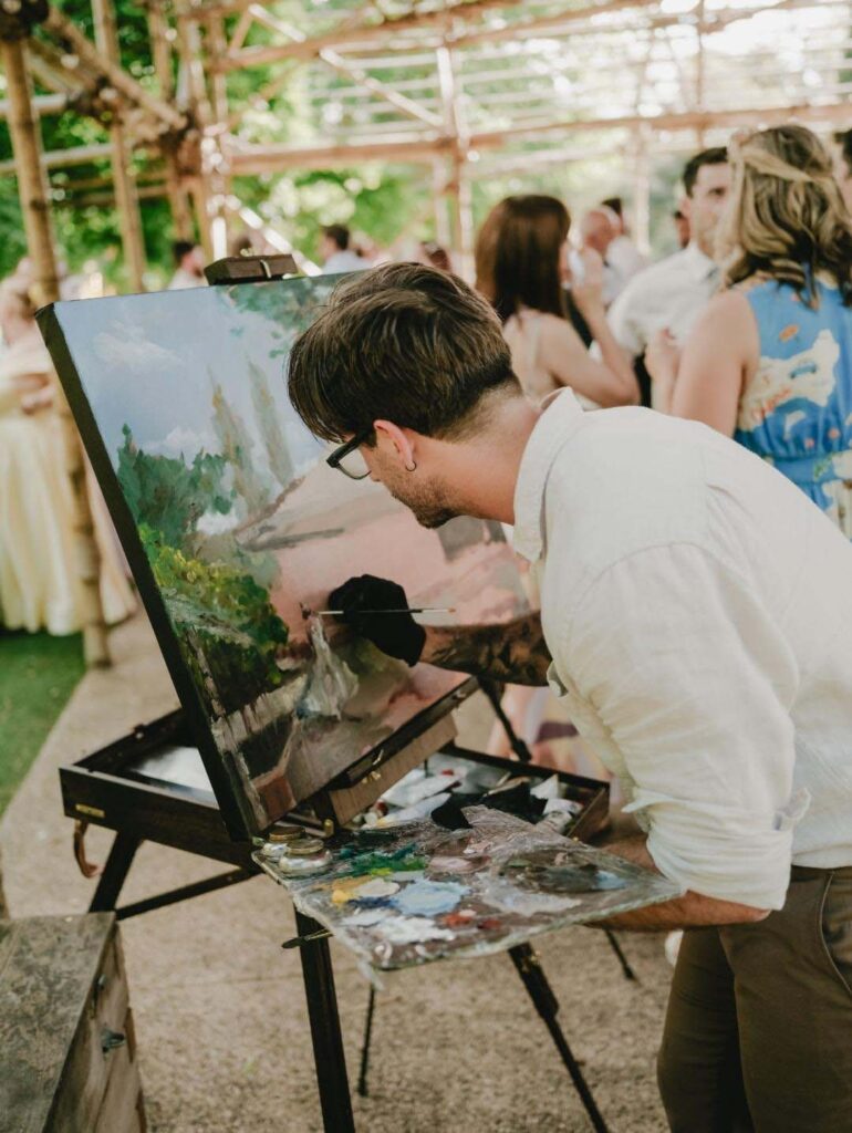 IMG_20251125_192505_496 | I do I do Gippsland Man in a white shirt and glasses painting on an outdoor easel at a garden party, with guests in the background.