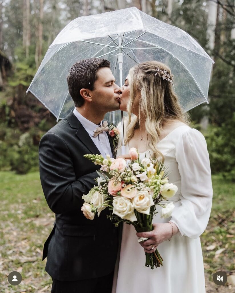 KATELAFF1 - I do I do Gippsland Bride and groom kissing under a clear umbrella in the rain, the bride holding a bouquet of pale pink and white flowers.
