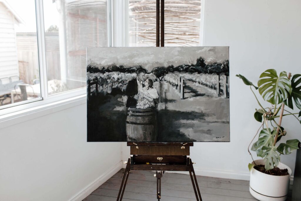 Kewish-13 | I do I do Gippsland Black-and-white painting of a couple embracing in a park, on an easel in a bright studio, with a potted plant to the right.