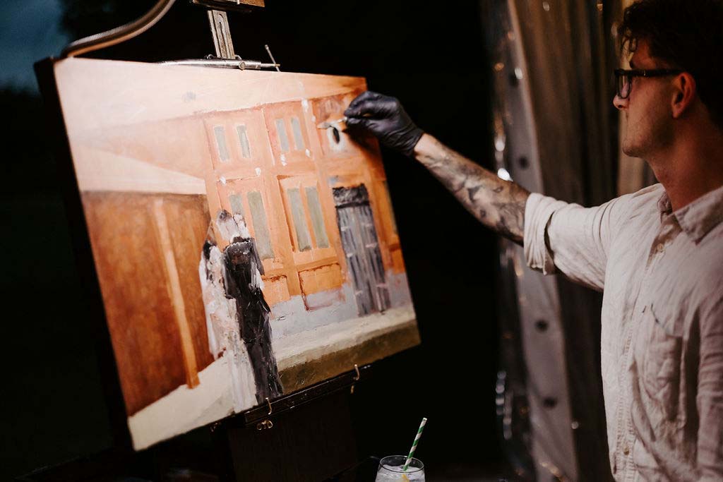 Kewish1 | I do I do Gippsland Artist wearing gloves and glasses painting a city street scene on a wooden canvas on an easel in a dim studio space.