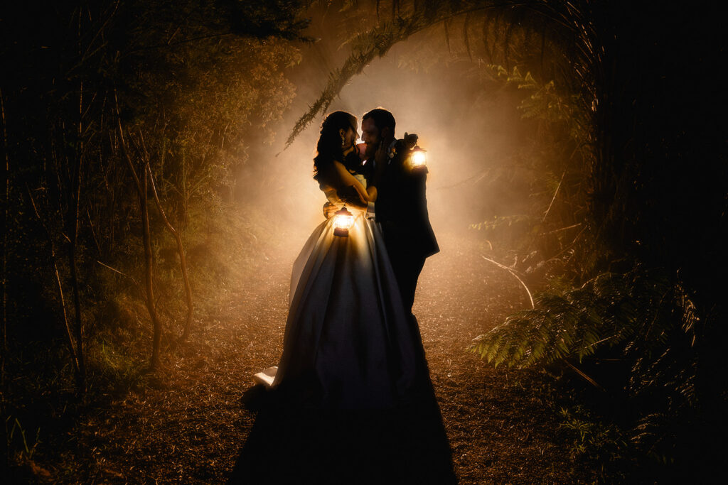 Mel and Riley - I do I do Gippsland Bride and groom embrace and kiss in a dark forest, lanterns casting warm light around them.