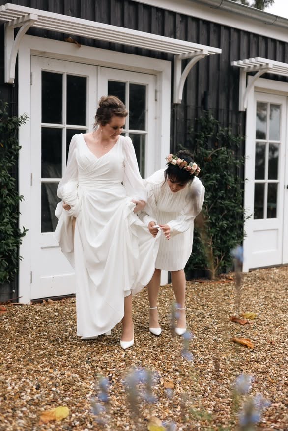 Molly Heath Photography 3 - I do I do Gippsland Bride in a white wedding gown is assisted by a bridesmaid in a white dress with a flower crown, as they walk on a gravel path outside a black wooden building.