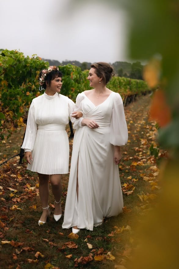 Molly Heath Photography1 - I do I do Gippsland Two women in white dresses walk arm in arm down a vineyard path, autumn leaves on the ground and a flower crown on one woman's head.