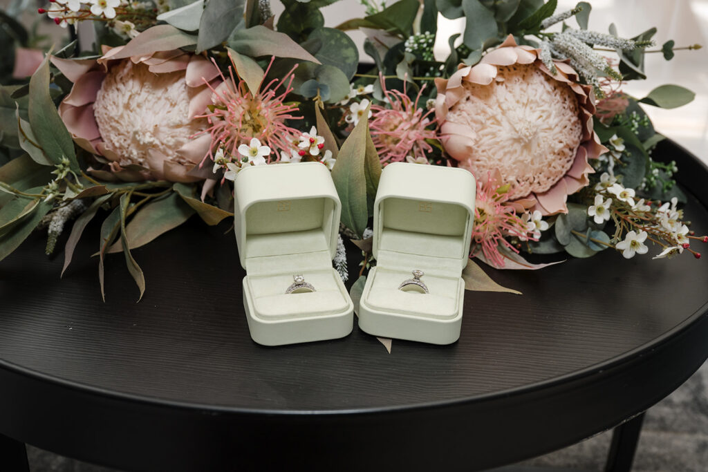 Two diamond engagement rings in pale green velvet boxes on a dark table amid a pink protea bouquet and greenery.