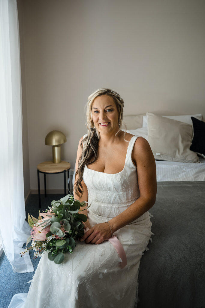 Bride in a white wedding gown sitting on a bed, holding a bouquet of greenery and pink flowers, smiling at the camera.