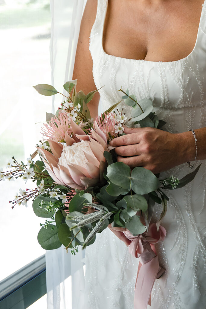Bride in a beaded white wedding dress holds a pink protea bouquet with eucalyptus, white blossoms, and a pink ribbon by a window.