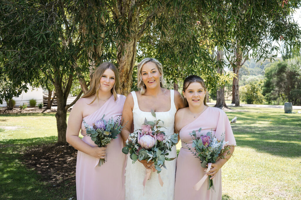 Bride in a white wedding dress stands between two bridesmaids in blush pink gowns, all smiling and holding pink and white bouquet arrangements under a leafy tree.