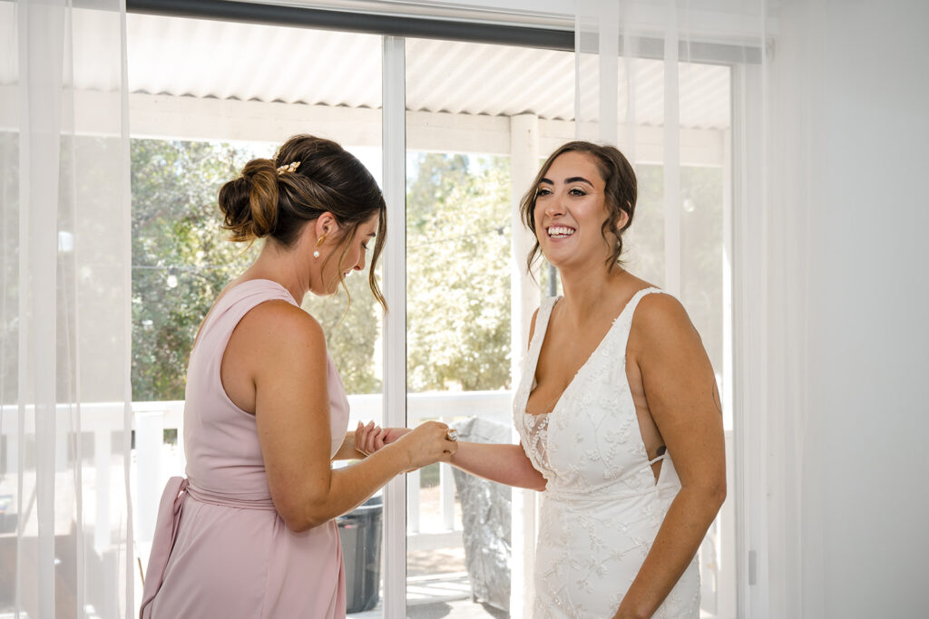Bride in a white lace wedding dress smiles as her bridesmaid in a blush pink dress helps fasten a bracelet on her wrist.