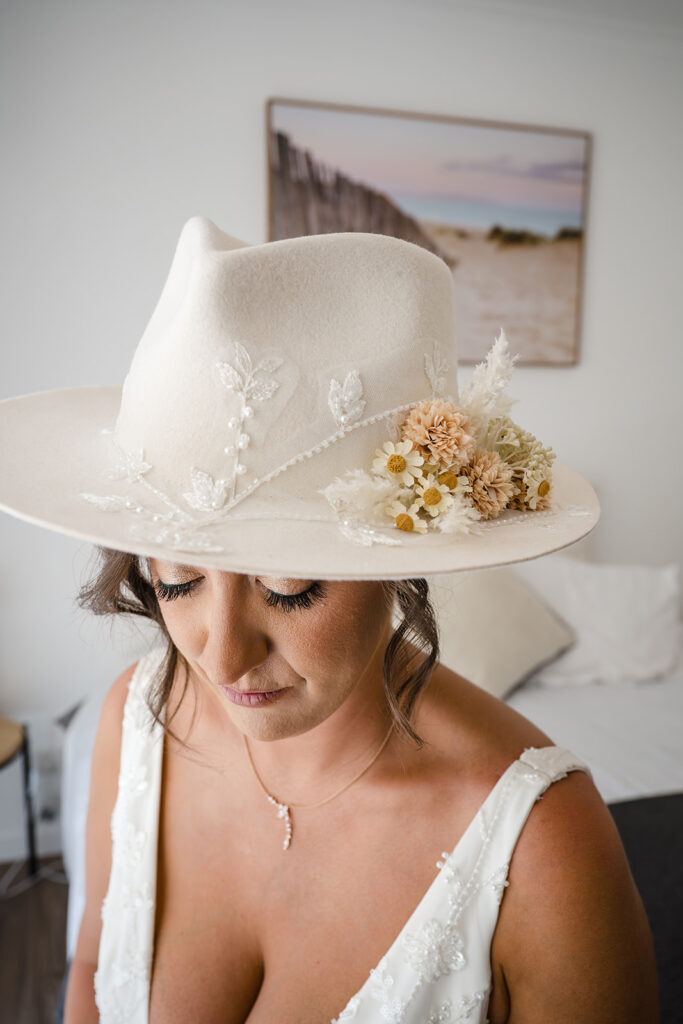 Bride wearing a wide-brim ivory hat with delicate lace and flower embellishments, matching a white wedding dress in a softly lit room.